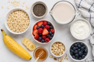 Overhead flat-lay of overnight oats ingredients including rolled oats, chia seeds, almond milk, Greek yogurt, fresh berries, banana, and honey arranged on a white kitchen surface